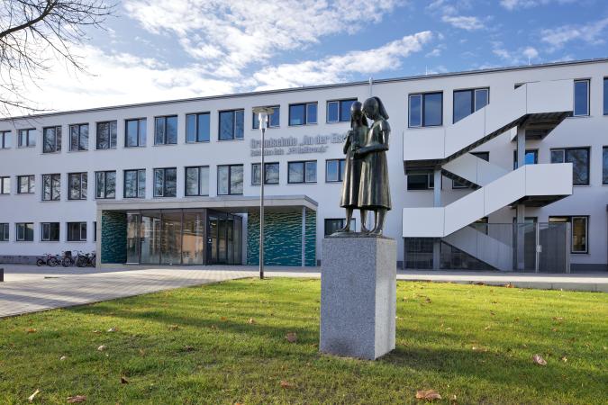Blick auf das Schulgebäude Haupteingang, im Vordergrund eine Statur mit zwei Mädchen