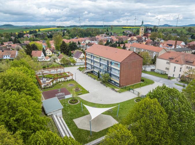 Blick auf das Schulgebäude von oben, im Vordergrund grüne Bäume, im Hintergrund Blick in die Weite