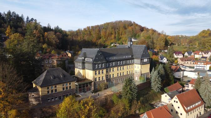 Blick auf das Schulgebäude mit Sportplatz, umgeben von herbstlichen Bäumen.