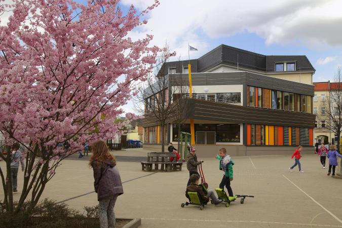 Blick auf das Schulgebäude, blühender Baum links, spielende Kinder auf dem Schulhof